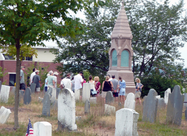 a group of people surround the tall pink granite mounment in the cemetery with gravestones in the foreground