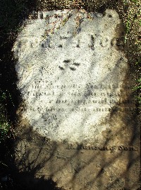 old marble grave marker fragment buried in the dirt with the shadow of a chain link fence over it