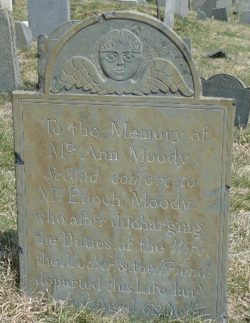old slate gravestone among other stones in a grave yard carved with a winged face and an inscription