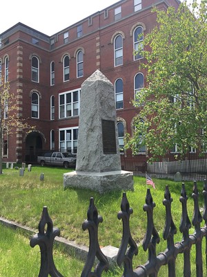 Granite pillar monument on top of a raised grassy square, bordered by granite curbing. The red bricked North School building is in the background. A leafy green tree is to the side and part of a fancy iron fence is in the foreground.