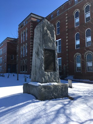 Granite pillar monument on top of a raised snowy berm. The red bricked North School building is in the background