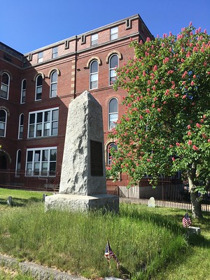 Granite pillar monument on top of a raised grassy square, bordered by granite curbing. The red bricked North School building is in the background. A leafy green tree with pink blossoms is shown to the side.